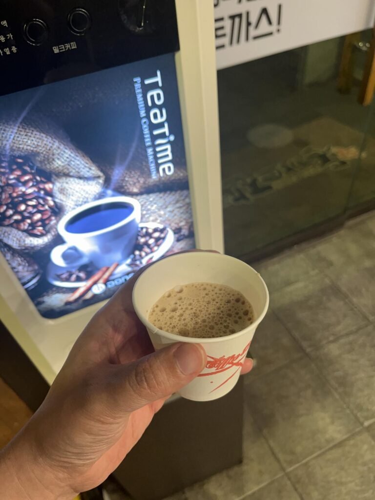 A hand holding a small paper cup of Korean mix coffee from a free vending machine at a restaurant entrance.