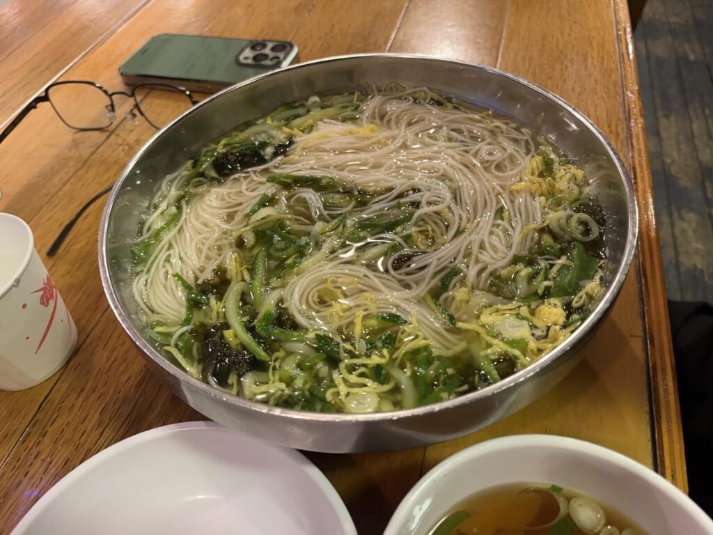 A stainless steel bowl of janchi-guksu, Korean banquet noodles in a clear broth topped with zucchini, egg, and seaweed.