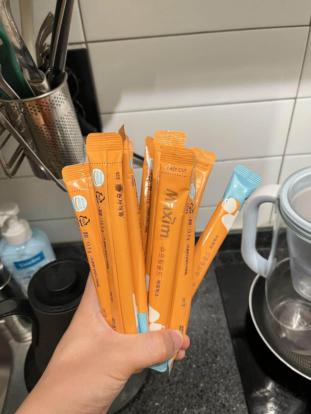 A hand holding a fan of orange Maxim Supreme Gold Korean coffee mix sticks in a kitchen.