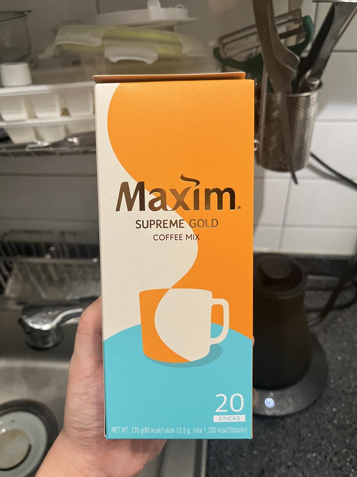 A hand holding a 20-stick box of Maxim Supreme Gold Korean coffee mix.