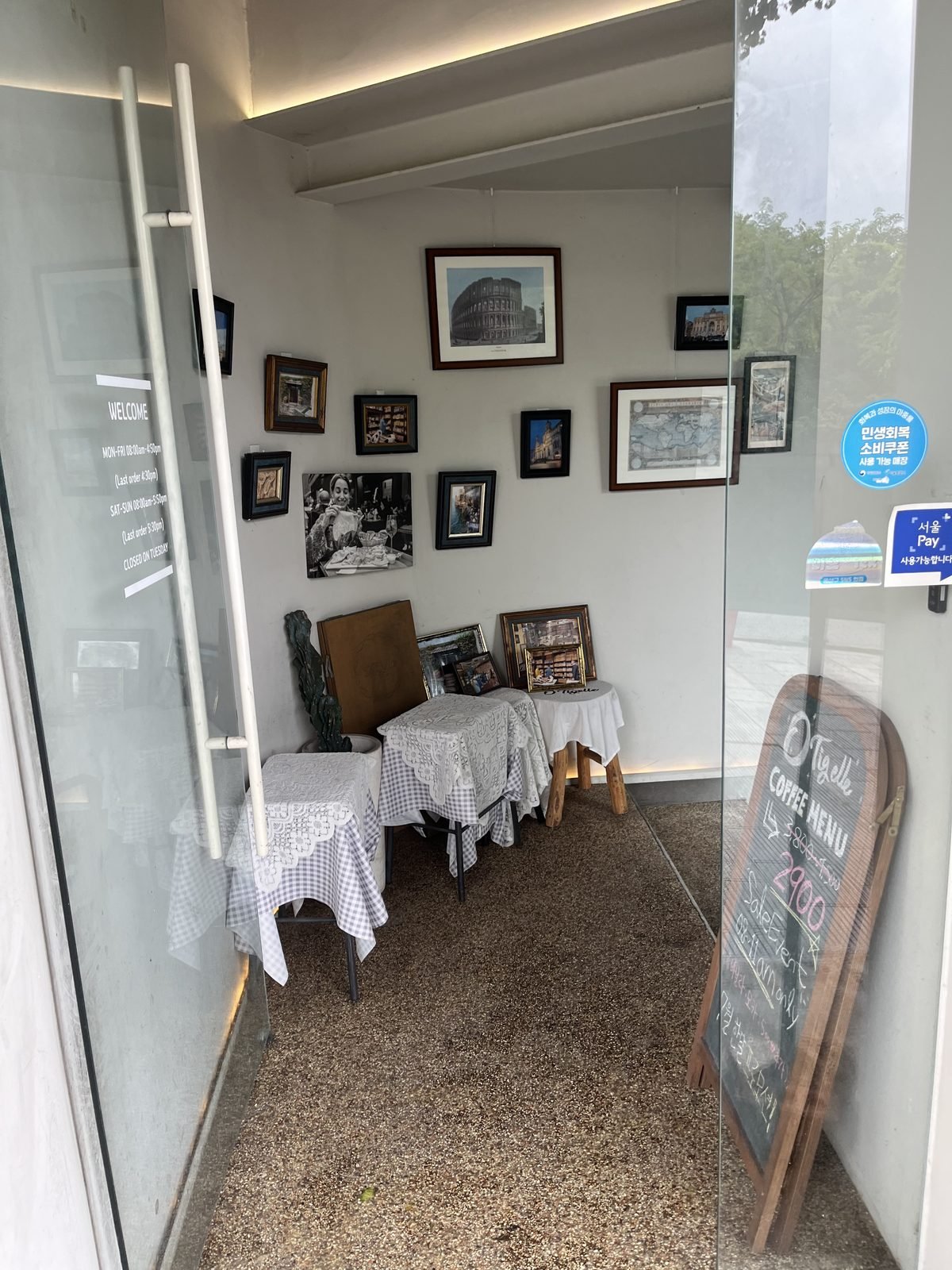 O Tigelle's glass entrance in Itaewon with opening hours on the door and framed prints of Italy inside