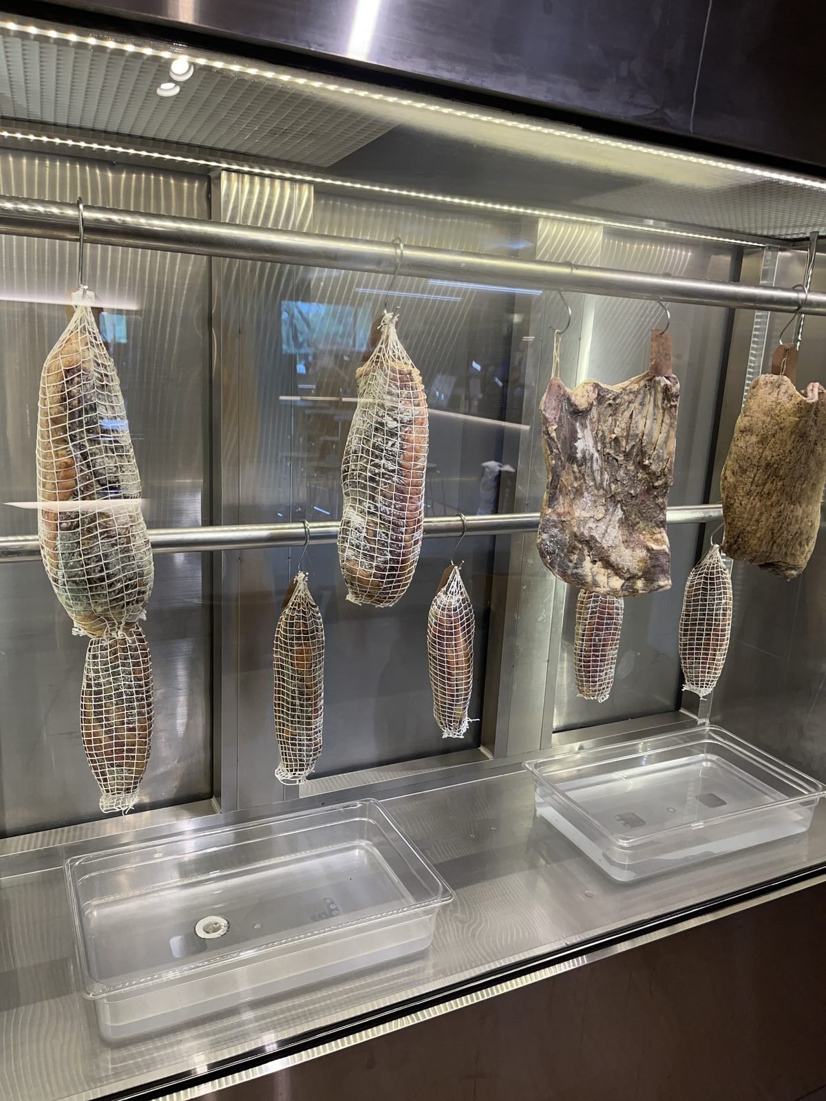 Whole cured meats in netting hanging in a glass curing cabinet at O Tigelle