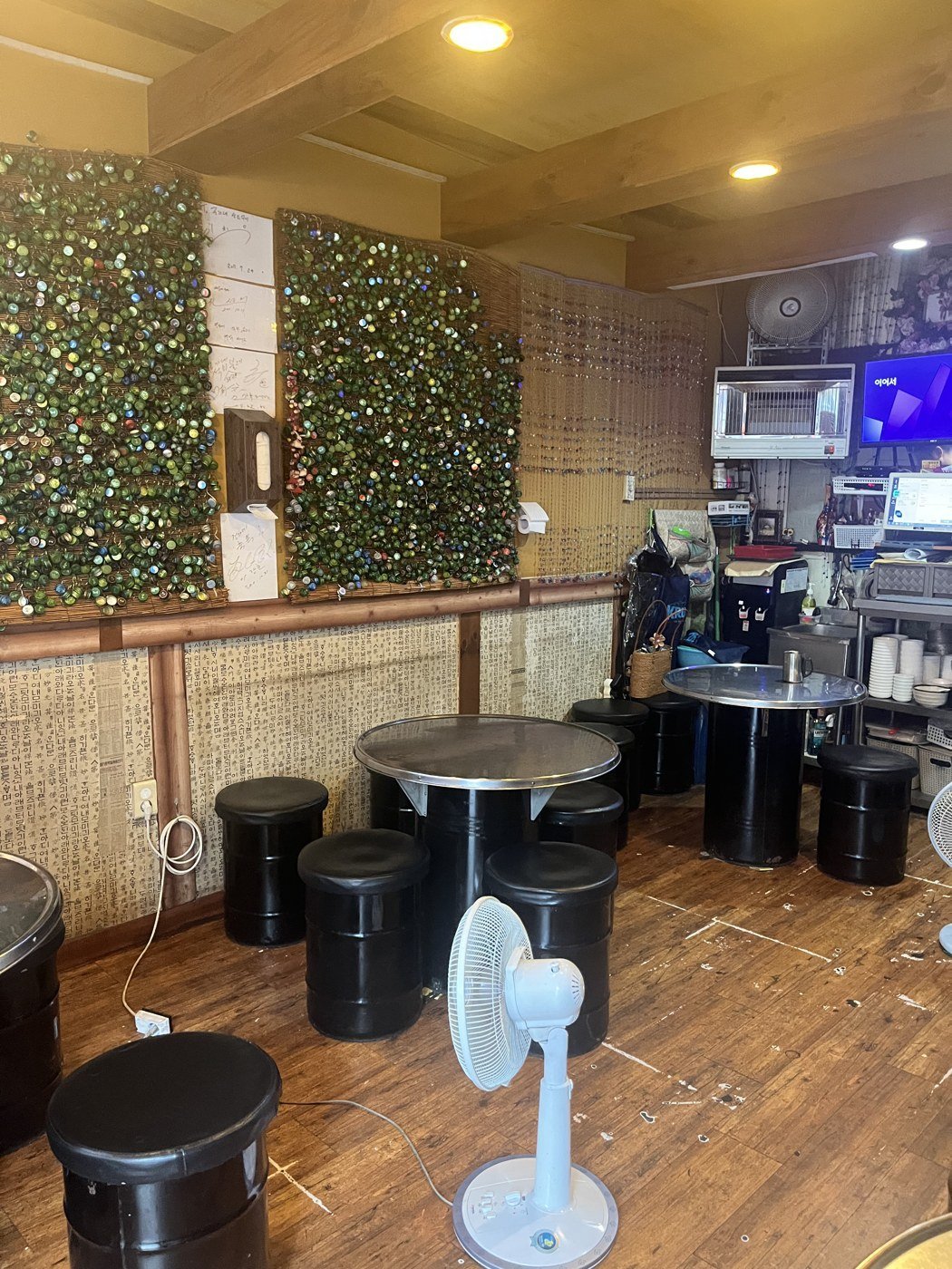 Inside: drum-can tables, black stools and a standing fan against a green wall.